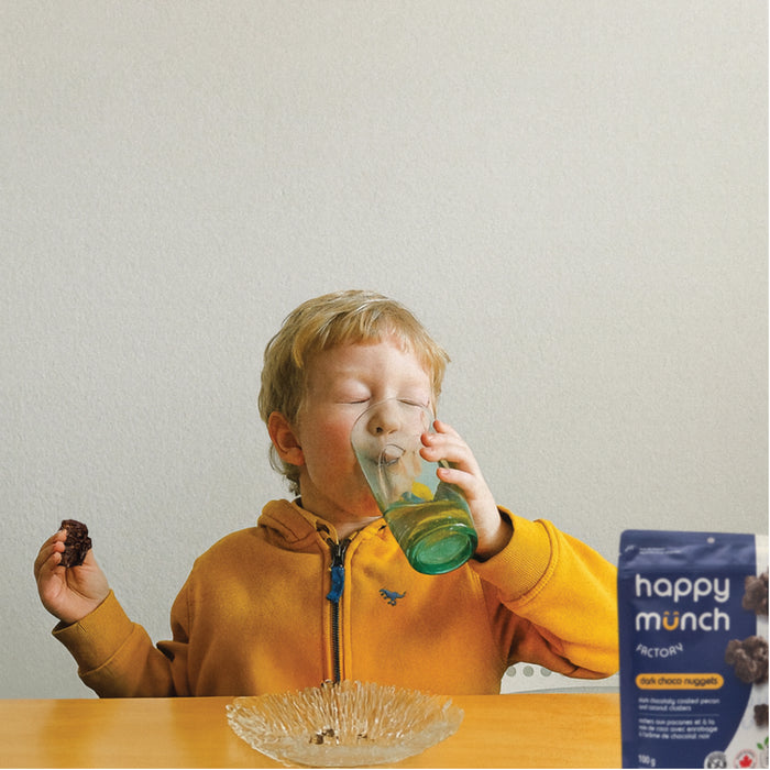 Child in a yellow jacket drinking from a green cup with a 'happy munch dark choco nuggets' pouch in the foreground.