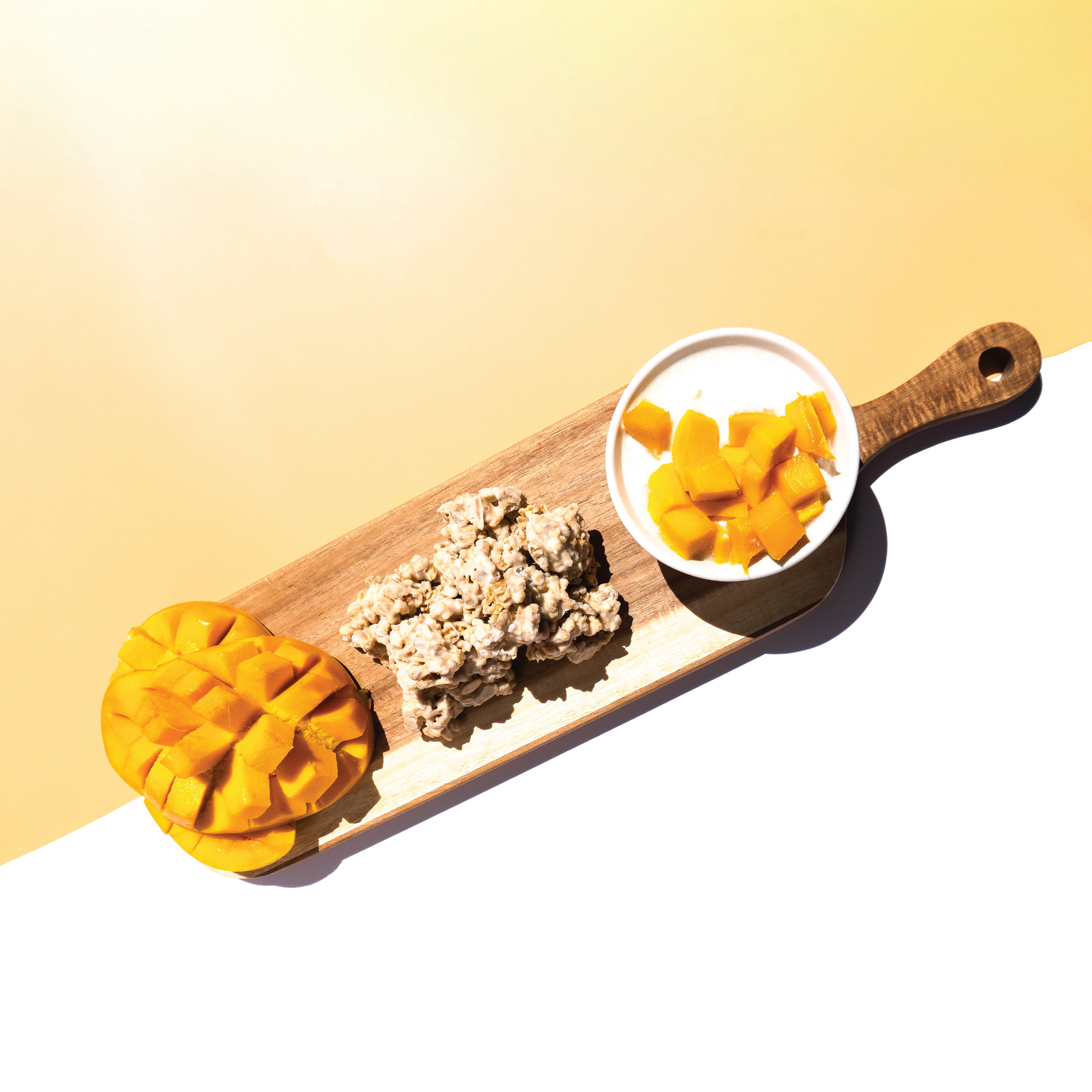 Wooden board with a bowl of mango and yogurt, a Happy Munch Mango Mania Yogurt Clusters, and a cut up mango on a gradient background.